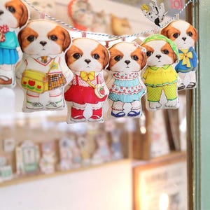 Pode incluir: Uma corda de enfeites de cachorro de tecido coloridos com diferentes roupas. Os enfeites est&atilde;o pendurados em uma corda branca. Os enfeites usam todos roupas diferentes, incluindo um uniforme escolar, um vestido vermelho, um vestido azul, uma camisa amarela e cal&ccedil;as, e uma camisa azul e cal&ccedil;as.
