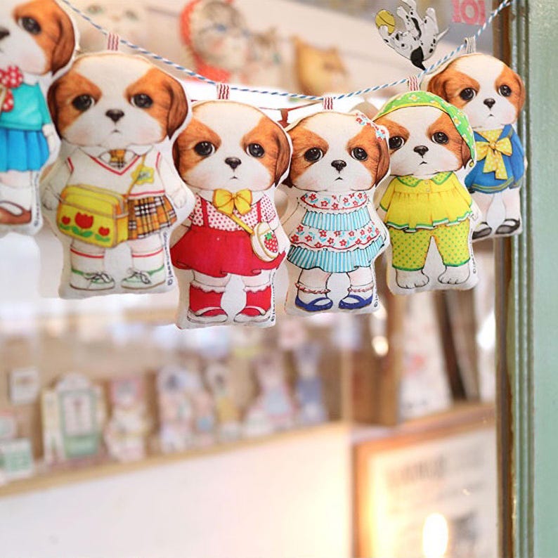 Pode incluir: Uma corda de enfeites de cachorro de tecido coloridos com diferentes roupas. Os enfeites est&atilde;o pendurados em uma corda branca. Os enfeites usam todos roupas diferentes, incluindo um uniforme escolar, um vestido vermelho, um vestido azul, uma camisa amarela e cal&ccedil;as, e uma camisa azul e cal&ccedil;as.