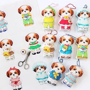 Pode incluir: Uma cole&ccedil;&atilde;o de 12 enfeites de pel&uacute;cia de cachorro, cada um vestido com uma roupa diferente. Os enfeites s&atilde;o todos brancos com orelhas e rostos marrons, e usam uma variedade de roupas coloridas, incluindo vestidos, camisas e cal&ccedil;as. Os enfeites est&atilde;o todos pendurados em uma corda.