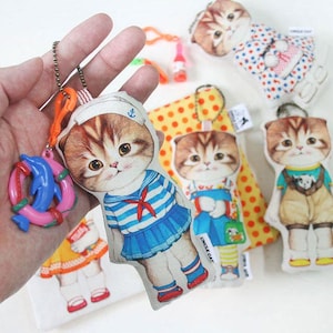 May include: A collection of fabric cat keychains featuring illustrations of kittens in various outfits, including a sailor suit, overalls, and polka dots. Each keychain has a metal ball chain and a small plastic charm, such as a life preserver.