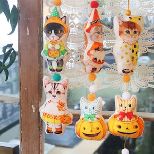 May include: A set of six Halloween-themed cat decorations. Each decoration features a different cat dressed in a costume, holding a pumpkin, or sitting inside a pumpkin. The decorations are made of fabric and have a vintage look.