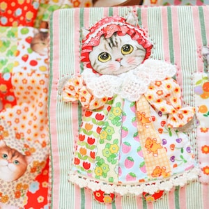 Op de afbeelding: Een patchwork-stofpop in de vorm van een kat met een roze-wit gestreepte jurk, een witte kantkraag en een roze-witte bloemenhoed. De pop zit op een roze-groene gestreepte stoffen achtergrond.