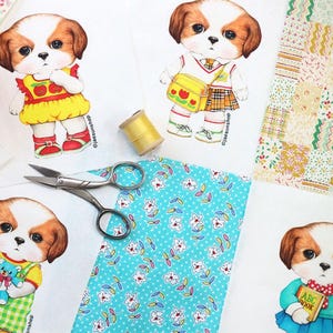 Pode incluir: Quatro quadrados de tecido impressos com ilustra&ccedil;&otilde;es de c&atilde;es vestindo roupas diferentes. Os c&atilde;es est&atilde;o vestindo um vestido amarelo com ma&ccedil;&atilde;s vermelhas, uma camisa branca com uma bolsa amarela, um vestido azul com um urso de pel&uacute;cia e um vestido verde com um livro. Os quadrados de tecido s&atilde;o cercados por tesoura, linha e um peda&ccedil;o de tecido com um padr&atilde;o floral.