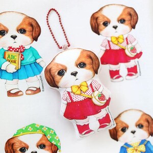 Pode incluir: Um chaveiro de peluche de um c&atilde;o que usa um vestido vermelho com bolinhas brancas, um la&ccedil;o amarelo e uma bolsa vermelha e branca em forma de morango. O c&atilde;o tem p&ecirc;lo castanho e grandes olhos castanhos. O chaveiro est&aacute; preso a uma corrente vermelha.