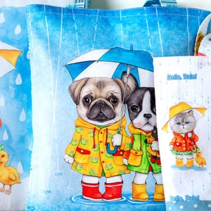 Könnte beinhalten: Eine blaue Tragetasche mit einer skurrilen Illustration von zwei Hunden in Regenmänteln, die einen Regenschirm halten. Die Hunde sind ein Mops und eine Französische Bulldogge. Der Hintergrund ist ein blauer Himmel mit Regentropfen. Der Text "Hello, Rain!" ist auf die Tasche gedruckt.