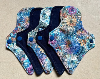 Puede incluir: Cinco compresas menstruales de tela reutilizables. Cada compresa presenta un vibrante estampado floral en tonos azules, morados, naranjas y blancos. Las compresas tienen un reverso azul oscuro y se aseguran con un cierre de botón a presión azul. Para la higiene femenina.
