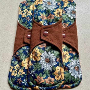 以下が含まれることがあります： 再利用可能な布製生理用ナプキン3枚。各ナプキンは、白、黄色、オレンジの花が咲く濃い青色の花柄です。茶色のボタン留めが付いています。快適さと環境に配慮したデザインです。