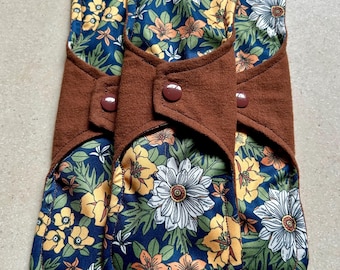 May include: Three reusable cloth menstrual pads. Each pad features a dark blue floral pattern with white, yellow, and orange flowers. The pads have a brown, buttoned closure. The pads are designed for comfort and eco-friendliness.