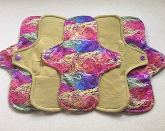 Puede incluir: Cinco compresas menstruales de tela reutilizables. Cada compresa presenta un colorido diseño arremolinado en tonos morado, rosa, naranja y verde en la capa superior. El núcleo absorbente es de color beige liso y cada compresa tiene un cierre de botón a presión.