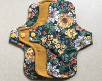 May include: Two reusable cloth menstrual pads with a floral print. The pads are layered with a dark blue fabric with a yellow floral pattern and a solid mustard yellow fabric.