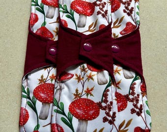 May include: Three reusable cloth menstrual pads with a white background and a pattern of red and white mushrooms, green leaves, and brown branches. The pads have a burgundy colored flannel lining.