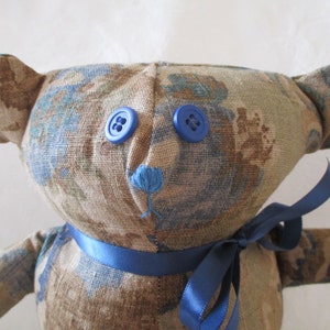 May include: Handmade teddy bear with a floral pattern in shades of blue, brown, and beige. The bear has blue button eyes, a blue embroidered nose, and a blue satin ribbon tied around its neck. A charming, handcrafted plush toy.