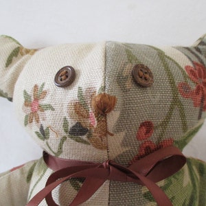 May include: A beige and green fabric teddy bear with brown buttons for eyes and a brown fabric bow tied around its neck.