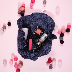 May include: A blue drawstring bag with white stars printed on it, filled with makeup products, including a pink blush labeled "Fit Me!" and a clear bottle of foundation. The bag is surrounded by small, colorful felt balls on a pink background.