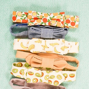 Könnte beinhalten: Fünf Stoff-Haarband mit verschiedenen Frucht-Prints. Die Haarband sind übereinander gestapelt, wobei das oberste Haarband Orangenscheiben zeigt, gefolgt von einem Jeans-Haarband, einem Haarband mit Bananen-Prints, einem Haarband mit Kiwischeiben und einem einfarbigen rosa Haarband.