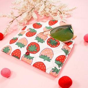 May include: A white quilted fabric case with a strawberry print and a green button closure. A pair of green sunglasses is partially inside the case.