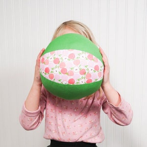 May include: A young person wearing a pink shirt with a floral pattern and a gray tiered skirt holds a green and pink ball with a peach pattern in front of their face.