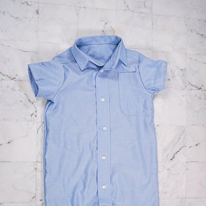 Op de afbeelding: Een lichtblauwe, kortgeknipte, knoopsluiting romper met een zakje aan de linkerkant.