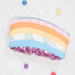 May include: A rainbow-colored zippered pouch with a floral pattern on the bottom. The pouch is surrounded by colorful pom-poms.