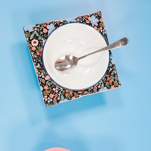May include: Two white ceramic bowls with spoons on top of floral patterned fabric placemats. The placemats are square and have a black background with colourful flowers. The other placemat is pink with a white background and colourful flowers.