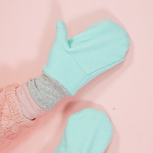 May include: A pair of light blue fleece mittens with grey cuffs. The mittens are shown against a pink background.