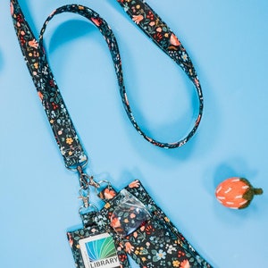 以下が含まれることがあります： 花柄の黒い紐と、黒い布製のバッジホルダーが2つ。1つのホルダーには透明なプラスチック製の窓があり、もう1つには図書館のカードが入っています。図書館のカードには、「LIBRARY」と「伝統、革新、多様性、包括性」の文字が書かれています。