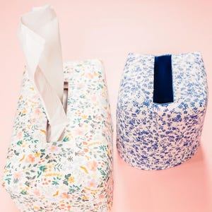 May include: Two fabric tissue box covers. One is rectangular with a floral pattern in pink, yellow, and green. The other is oval-shaped with a blue floral pattern and a dark blue opening. Both are on a pink background.
