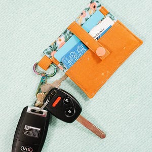 May include: Orange fabric cardholder with a floral patterned accent and a snap closure. Includes a keychain with keys and a car key fob. The cardholder holds several cards, including one that says "IS COUNTY".