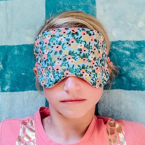May include: A person wearing a pink shirt with a gold giraffe print and a floral eye mask. The eye mask is blue with pink flowers and gold accents.