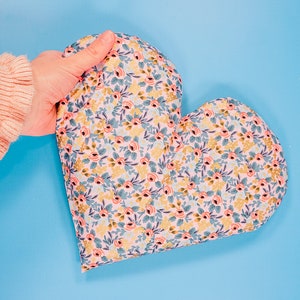 Può includere: Un cuscino a forma di cuore con un motivo floreale in tonalità rosa, blu e oro. Il cuscino è realizzato in tessuto e ha una texture morbida e soffice.