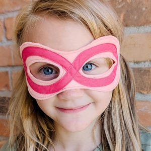 May include: A pink and fuchsia superhero mask with an infinity symbol design. The mask is made of soft fabric and is worn by a child with blonde hair and blue eyes. The mask is a fun accessory for dress-up and imaginative play.