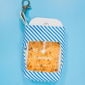 May include: A clear hand sanitizer spray bottle with the text "touchland" is encased in a blue and white striped fabric holder. The holder has a silver clip and a white top. The background is a solid light blue.