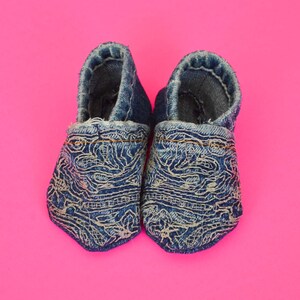 May include: A pair of denim baby shoes with a white embroidered pattern. The shoes are made of blue denim and have a white embroidered pattern on the front. The shoes are on a pink background.