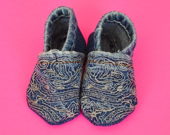 Könnte beinhalten: Ein Paar Baby-Schuhe aus Jeans mit einem weißen Stickmuster. Die Schuhe sind aus blauem Jeans und haben ein weißes Stickmuster auf der Vorderseite. Die Schuhe befinden sich auf einem rosa Hintergrund.