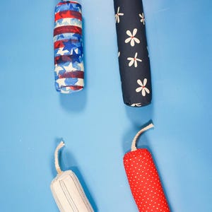 May include: Four fabric decorative items resembling firecrackers. One is red, white, and blue with stars and stripes. Another is navy blue with white flowers. The other two are white with blue stripes and red with white polka dots.