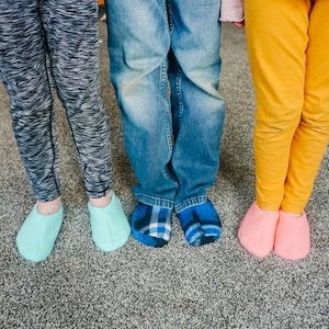 May include: Four pairs of feet wearing different colored slippers. The slippers are all soft and cozy, and they are perfect for keeping your feet warm in the winter. The feet are wearing a variety of socks, including plaid socks and solid colored socks.