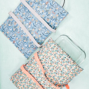 May include: Two floral patterned oven mitts with handles. The mitts are folded and a glass baking dish is placed on top of one of the mitts.