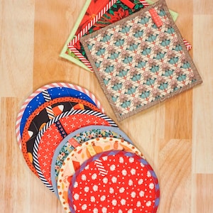 May include: A collection of quilted kitchen items including three square potholders with geometric, floral, and holiday patterns, and a stack of seven circular fabric coasters featuring various colorful prints like red daisies, orange pumpkins, and blue stars.