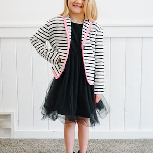 Peut inclure: Une jeune fille portant une robe en tulle noire avec un blazer rayé blanc et noir avec des bordures roses. Elle porte des bottines noires.