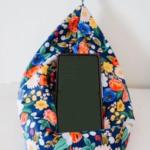 Op de afbeelding: Een blauwe en witte bloemen telefoonstandaard met een smartphone erin. De telefoonstandaard is kussenvormig en heeft een kleine lus aan de bovenkant.