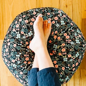 May include: A person's legs are crossed and resting on a round floral patterned bean bag chair. The bean bag is black with pink and green flowers. The person is wearing blue jeans.