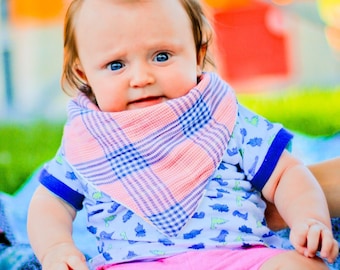May include: A baby with spiky blonde hair wearing a pink and blue plaid bandana and a blue shirt with a dinosaur print. The baby is sitting on a blue and white blanket.