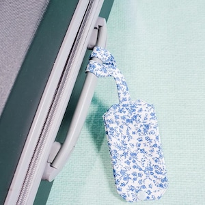 Puede incluir: Una etiqueta de equipaje blanca con un estampado floral azul está sujeta a un asa de maleta verde. La etiqueta es rectangular con bordes redondeados y un lazo para sujetar. El fondo es verde claro.