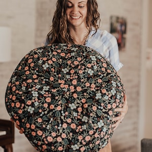 May include: A woman holds a large round black and white floral patterned cushion. The cushion has a dark background with pink and white flowers and green leaves.