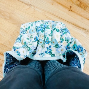 Può includere: Le gambe di una persona sono coperte da una coperta con un motivo floreale blu e bianco con un bordo blu chiaro. La coperta è piegata sulle gambe della persona e la persona indossa pantaloni blu scuro.