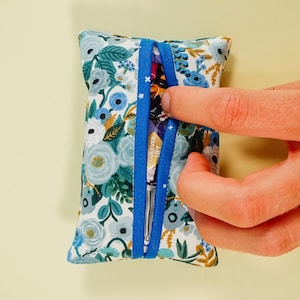 May include: A small, rectangular fabric pouch with a floral pattern in shades of blue, white, and gold. The pouch is accented with a blue ribbon and contains a packet. The pouch is being opened by a person.