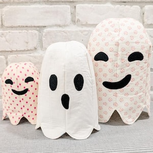 May include: Three fabric ghost-shaped decorations of varying sizes. The smallest and largest ghosts feature a white fabric with a repeating pink circular pattern and black felt eyes and mouths. The middle ghost is solid white with black felt eyes and mouth.