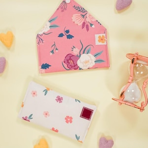 Op de afbeelding: Twee stoffen enveloppen met bloemenpatronen. De grotere envelop is roze met een wit bloemenpatroon. De kleinere envelop is wit met een roze bloemenpatroon. Beide enveloppen hebben een klein vierkant stuk stof in een contrasterende kleur. De enveloppen zijn omringd door kleine vilt hartjes in verschillende kleuren.