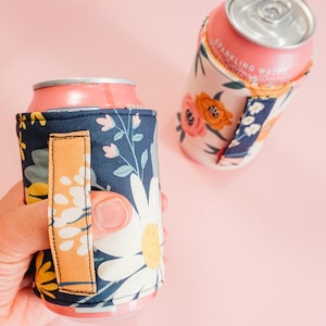 以下が含まれることがあります： 2つのフローラル柄の缶クーラー。1つは手で持っています。クーラーは、大きな白いデイジーと黄色のアクセントが付いた濃いネイビーブルーの背景が特徴です。もう1つのクーラーは、ピンクとオレンジの花が付いた白い背景です。