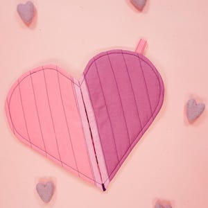 Puede incluir: Un agarrador de olla en forma de corazón rosa y morado con un patrón a rayas. El agarrador de olla está hecho de tela y tiene un lazo para colgar.